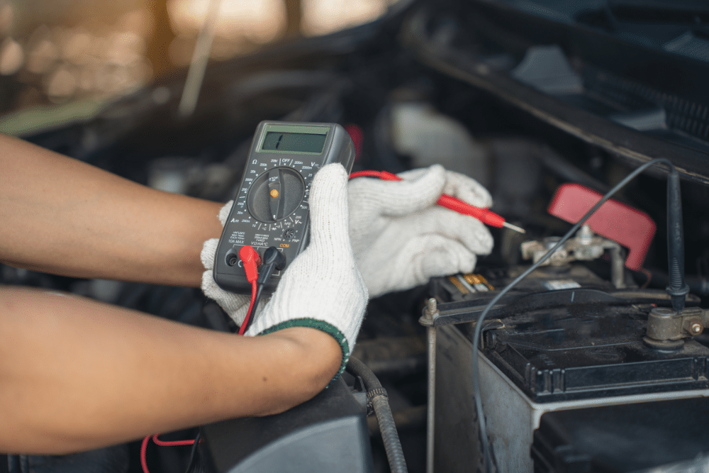 A person wearing white gloves uses a digital multimeter to check a car battery under the hood of a vehicle.