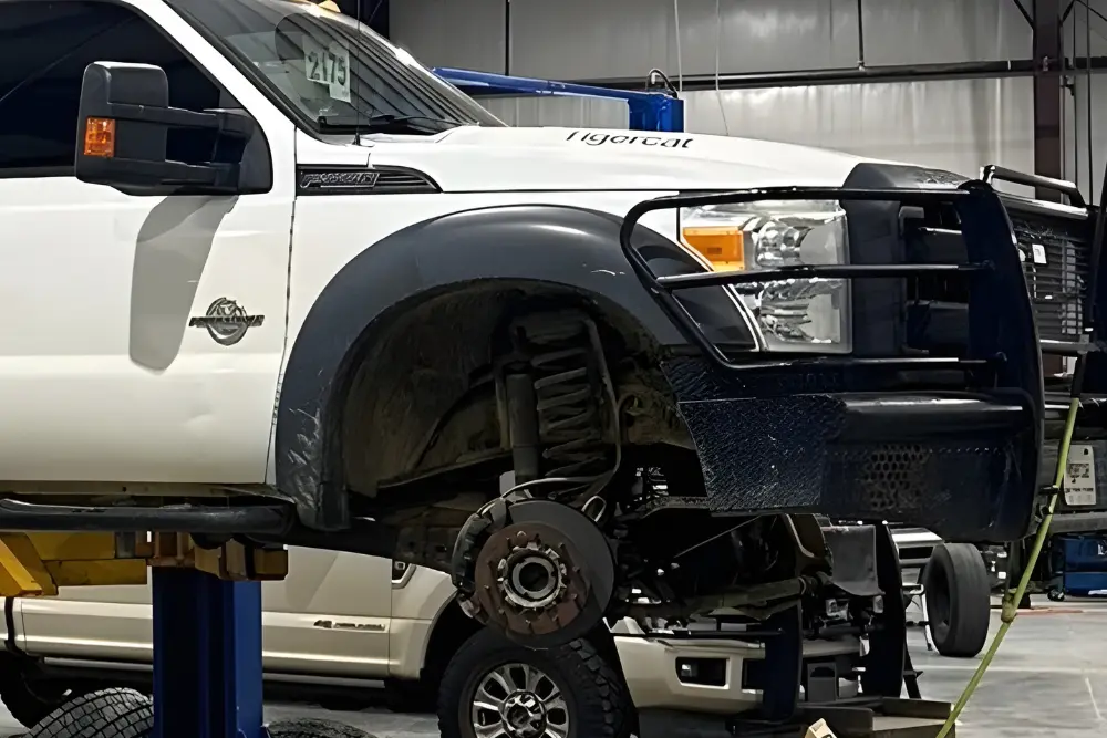 A white pickup truck is lifted on a hydraulic car lift inside a garage, with its front wheel removed, exposing the suspension and brake components. Another vehicle is visible in the background.