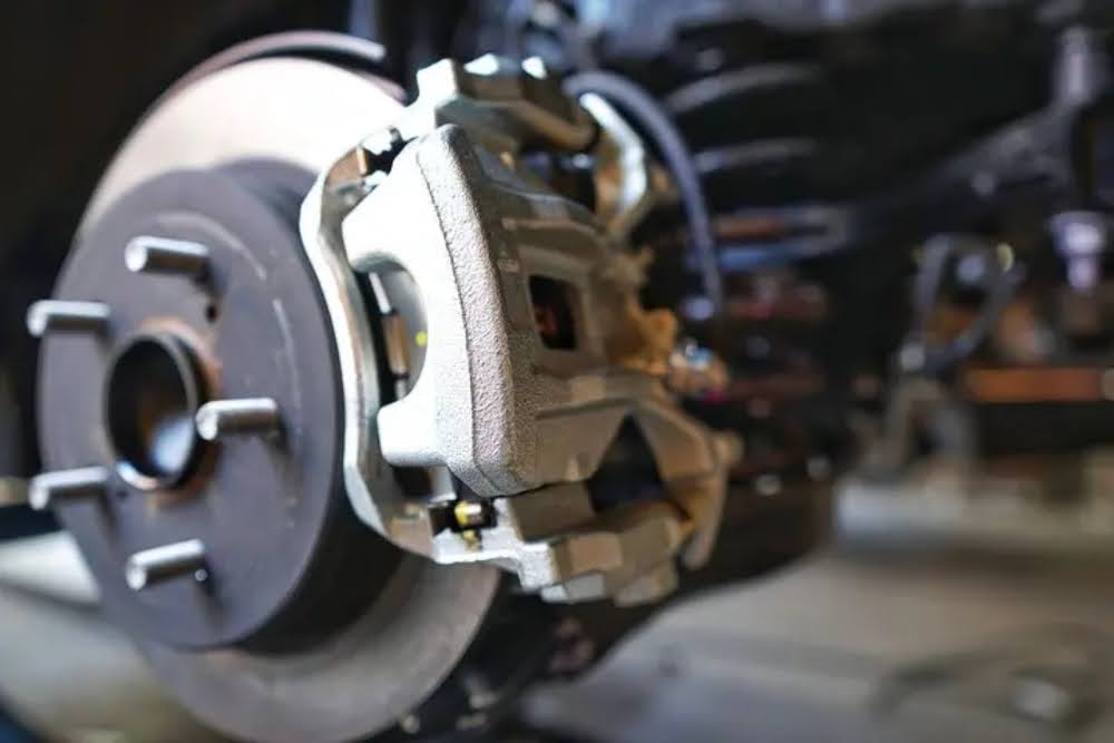 Close-up of a car’s disc brake assembly, showing the brake rotor and silver caliper, with some suspension components visible in the background.