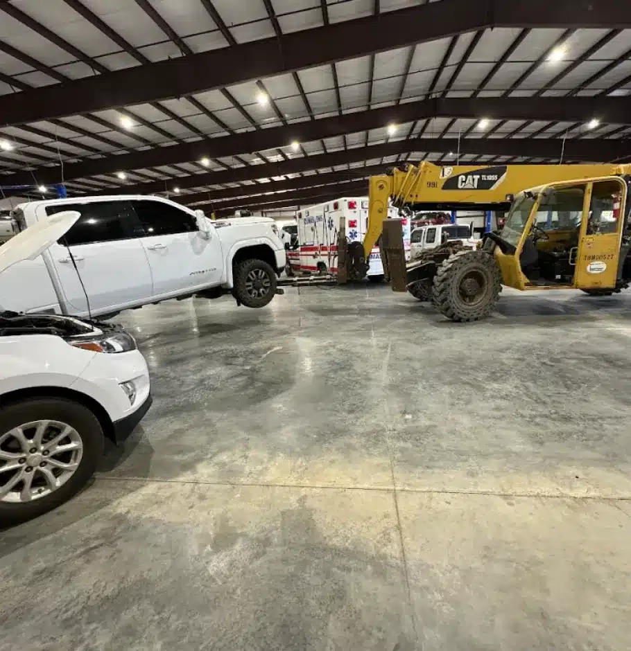 Diesel Repair In Monticello AR At Strokers Diesel Performance & Repair. A yellow CAT telehandler is lifting the rear end of a white pickup truck inside a large warehouse with a concrete floor; another white vehicle with its hood open is in the foreground.