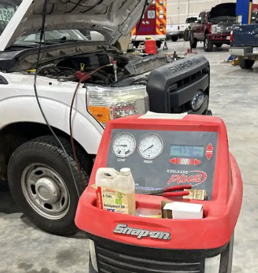 Auto Air Conditioning & Heater Repair In Monticello AR At Strokers Diesel Performance & Repair. A white Ford Super Duty truck with its hood open is being serviced indoors. In the foreground, a red Snap-on KoolKare Plus machine with gauges and tools is visible. Other vehicles and equipment are in the background.