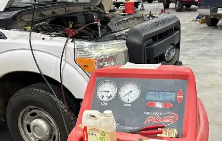 Auto Air Conditioning & Heater Repair In Monticello AR At Strokers Diesel Performance & Repair. A white Ford truck with its hood open is being serviced in a garage. In the foreground, an automotive coolant recovery machine with gauges and tools is visible.