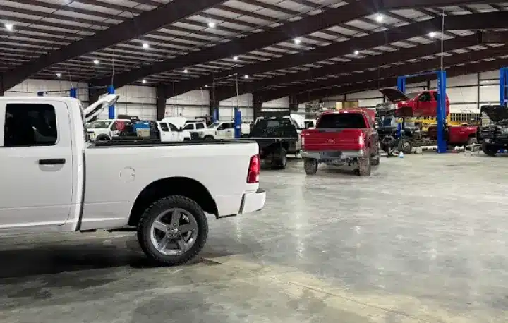 Auto Repair In Monticello AR At Strokers Diesel Performance & Repair. A spacious auto repair shop with multiple pickup trucks parked and lifted on blue hydraulic car lifts, under a large metal roof with bright overhead lights. The floor is clean and the area is well-organized.