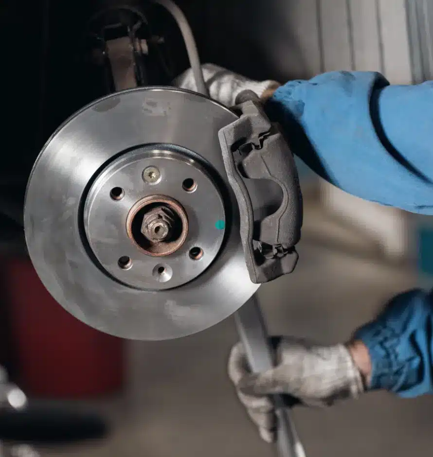 Brake Repair In Monticello AR At Strokers Diesel Performance & Repair. A person wearing gloves and a blue sleeve is working on a car’s exposed brake disc and caliper, using a tool to perform maintenance or repair.