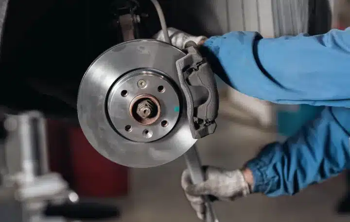 Brake Repair In Monticello AR At Strokers Diesel Performance & Repair. A mechanic in blue coveralls and gloves works on a cars brake disc and caliper, using a tool to perform maintenance or repairs in an automotive workshop.