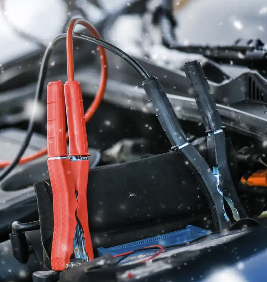 Auto Electrical Repair In Monticello AR At Strokers Diesel Performance & Repair. A close-up of red and black jumper cables connected to a car battery under the hood, with some snowflakes falling around the engine area.
