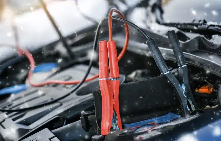 Electrical Repair In Monticello AR At Strokers Diesel Performance & Repair. Close-up of red and black jumper cables attached to a car battery under the hood, with snowflakes falling, suggesting a car jump start in cold winter weather.
