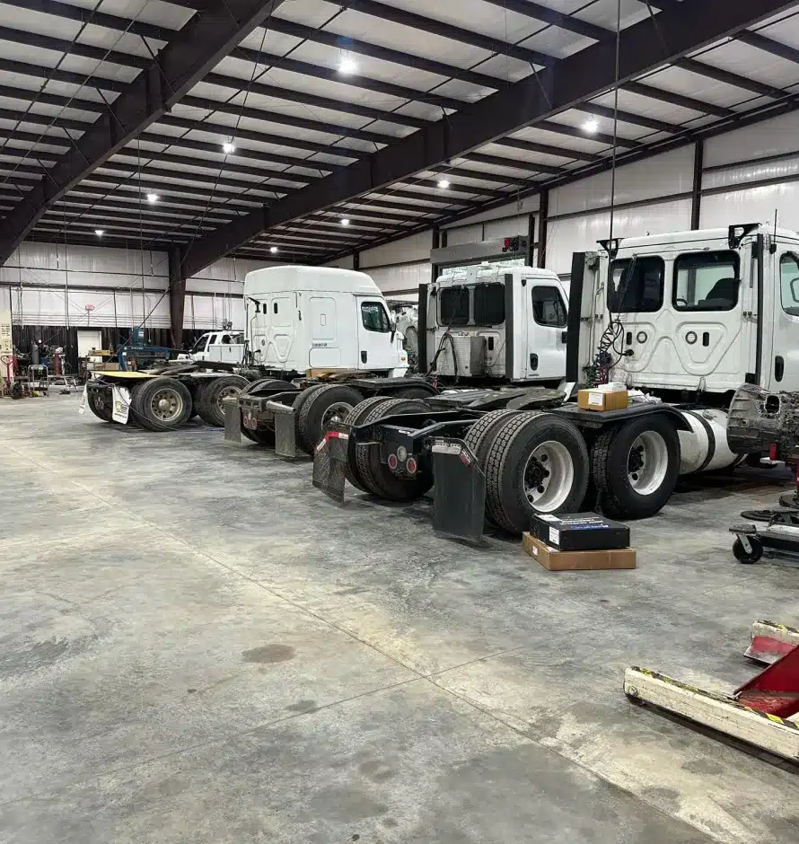 Fleet Maintenance In Monticello AR At Strokers Diesel Performance & Repair. Several large white semi-truck cabs are parked in a spacious, well-lit garage or warehouse, with various tools and equipment scattered on the concrete floor.