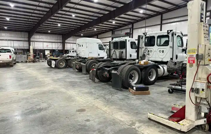Fleet Maintenance In Monticello AR At Strokers Diesel Performance & Repair. Several large white trucks without trailers are parked inside a spacious warehouse with high ceilings, exposed beams, and concrete floors. Various tools and equipment are scattered around the area.