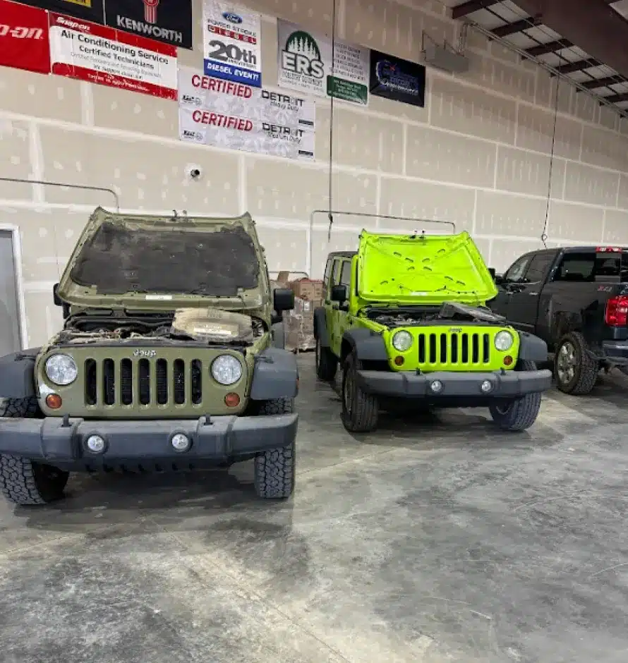 Vehicle Inspections In Monticello AR At Strokers Diesel Performance & Repair. Two green Jeep Wranglers with their hoods open are parked indoors side by side, next to a dark-colored pickup truck. The vehicles are in a garage or workshop with banners and unfinished walls in the background.