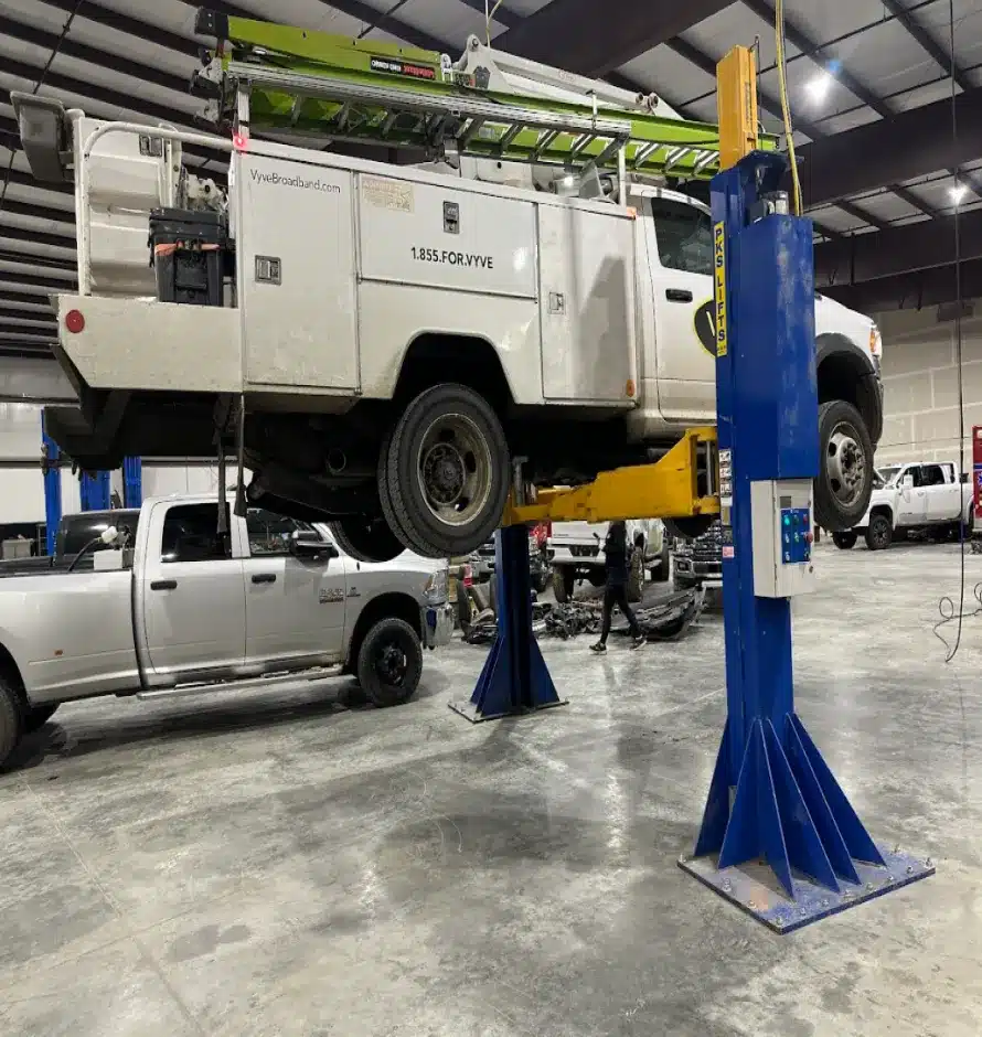 Preventative Maintenance & Oil Change In Monticello AR At Strokers Diesel Performance & Repair. A white utility truck is lifted by a blue hydraulic car lift in a spacious garage or workshop. Other vehicles and equipment are visible in the background on the concrete floor.