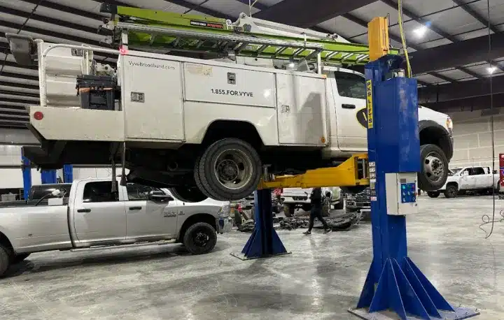 Preventative Maintenance & Oil Change In Monticello AR At Strokers Diesel Performance & Repair. A white utility truck is elevated on a hydraulic lift inside an auto repair shop. Another pickup truck and various equipment are visible in the spacious, well-lit garage.