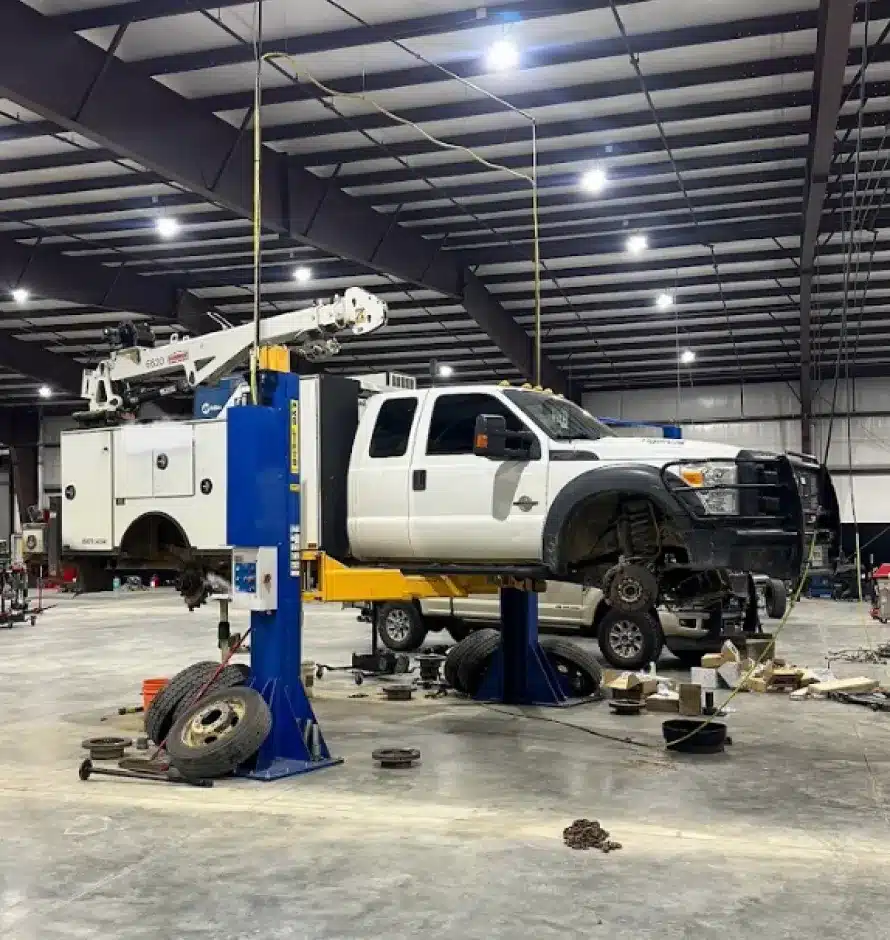 Suspension Repair In Monticello AR At Strokers Diesel Performance & Repair. A white utility truck is lifted on a blue hydraulic car lift inside a large garage or workshop. The trucks front wheels are removed, and various tools and tires are scattered on the floor nearby.