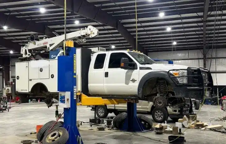 Suspension Repair In Monticello AR At Strokers Diesel Performance & Repair. A white utility truck is lifted on a hydraulic car lift inside a large garage, with its front wheels removed and tools and tires scattered on the floor around it.