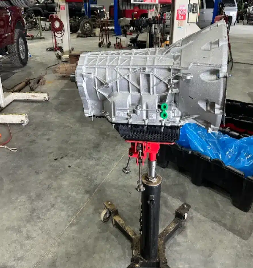 Transmission Repair & Replacement In Monticello AR At Strokers Diesel Performance & Repair. A car transmission mounted on a hydraulic jack stands in the middle of an automotive workshop, with various vehicles, tools, and equipment visible in the background.