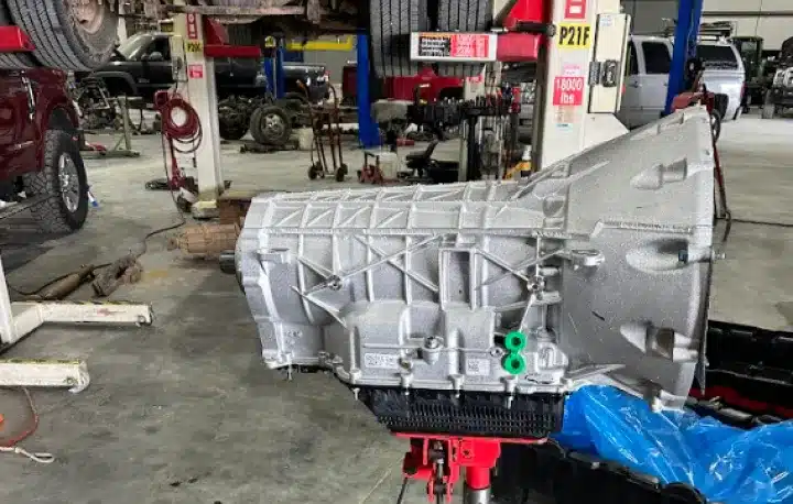 Transmission Services & Replacement In Monticello AR At Strokers Diesel Performance & Repair. A silver automotive transmission sits on a stand in a busy auto repair shop, with various vehicles, tools, and equipment visible in the background.