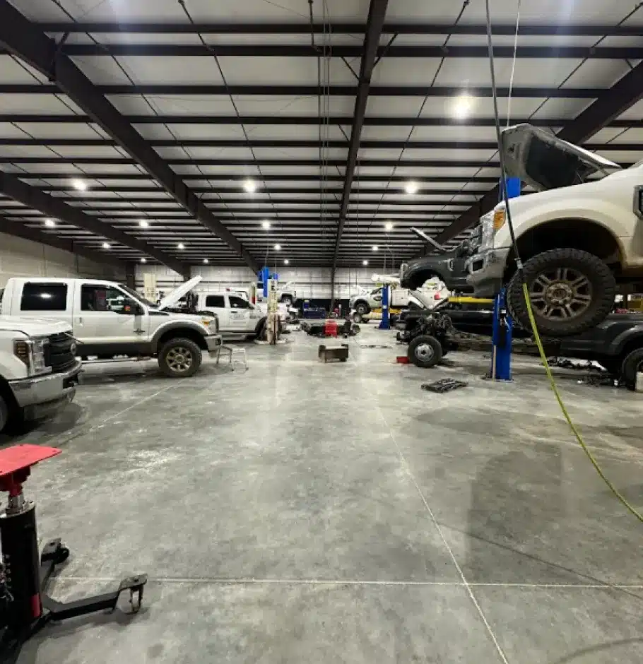 Vehicles We Service In Monticello AR At Strokers Diesel Performance & Repair. Large auto repair shop with multiple white pickup trucks being serviced. Some trucks are lifted on car lifts, hoods open, and tools or parts scattered around the spacious, well-lit workshop with a concrete floor.