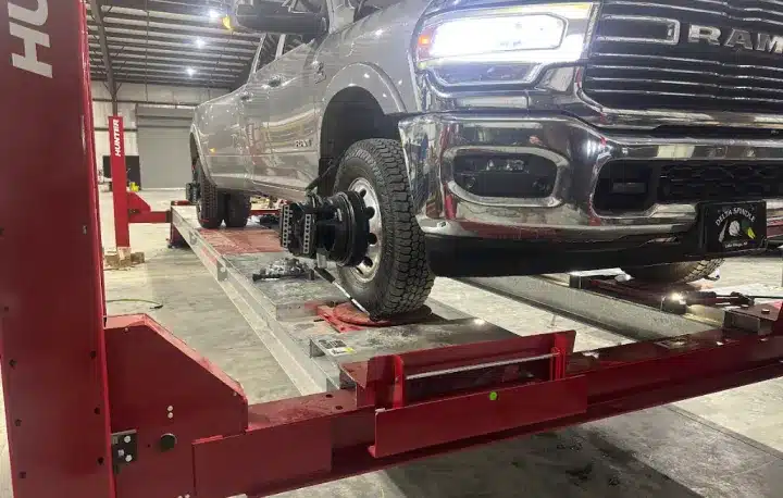 Wheel Alignment In Monticello AR At Strokers Diesel Performance & Repair. A pickup truck is raised on a red hydraulic lift inside a garage; its front passenger-side wheel has been removed, exposing the brake assembly.