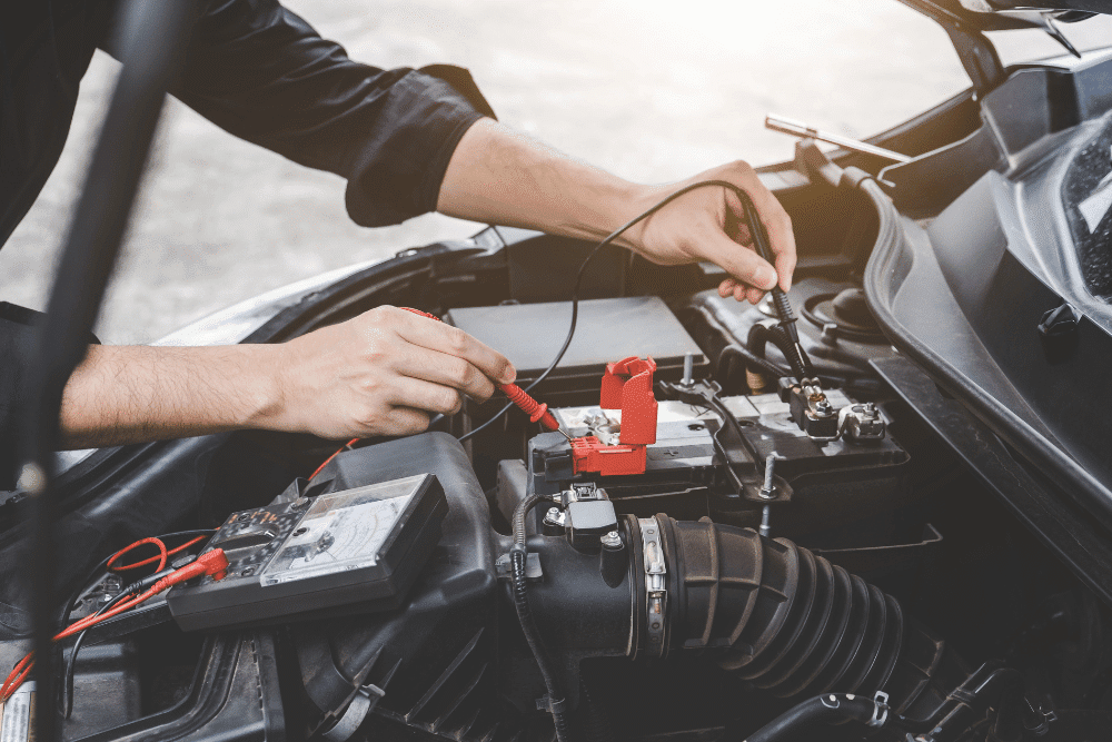 preventative maintenance during national car care month by Strokers Diesel Performance and Repair in Monticello, AR -- A person uses multimeter probes to check a car battery under the open hood of a vehicle.