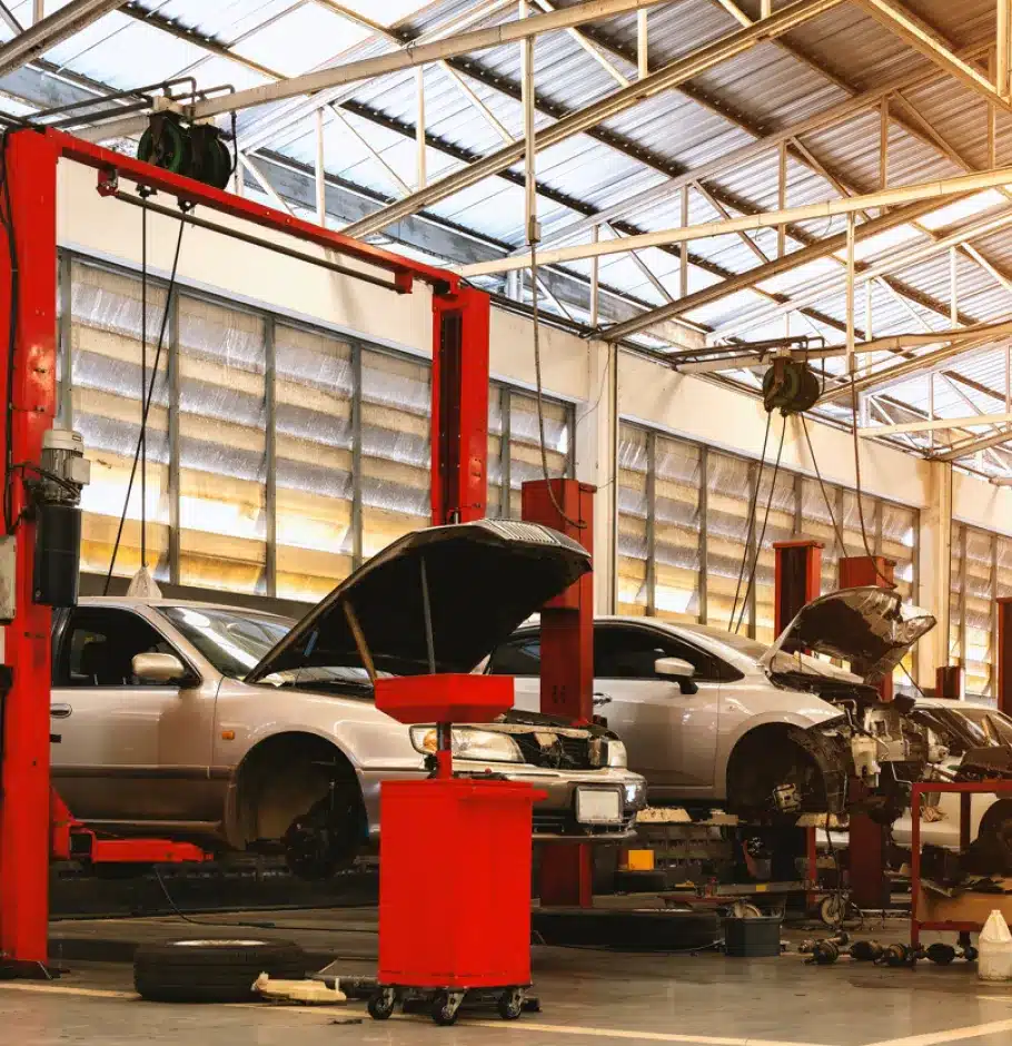 Asian Vehicle Repair In Monticello AR At Strokers Diesel Performance & Repair. Two cars with open hoods are parked inside a brightly lit auto repair shop. Red hydraulic lifts and various tools surround the vehicles, and sunlight streams through large windows above.