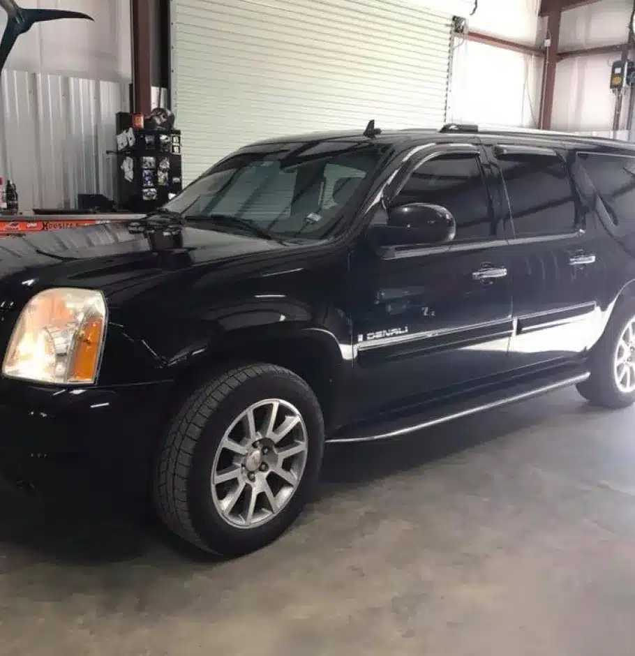 Domestic Vehicle Repair In Monticello AR At Strokers Diesel Performance & Repair. A black GMC Yukon Denali SUV is parked indoors on a concrete floor near a white roll-up garage door in a metal building. The vehicle has tinted windows and silver rims.