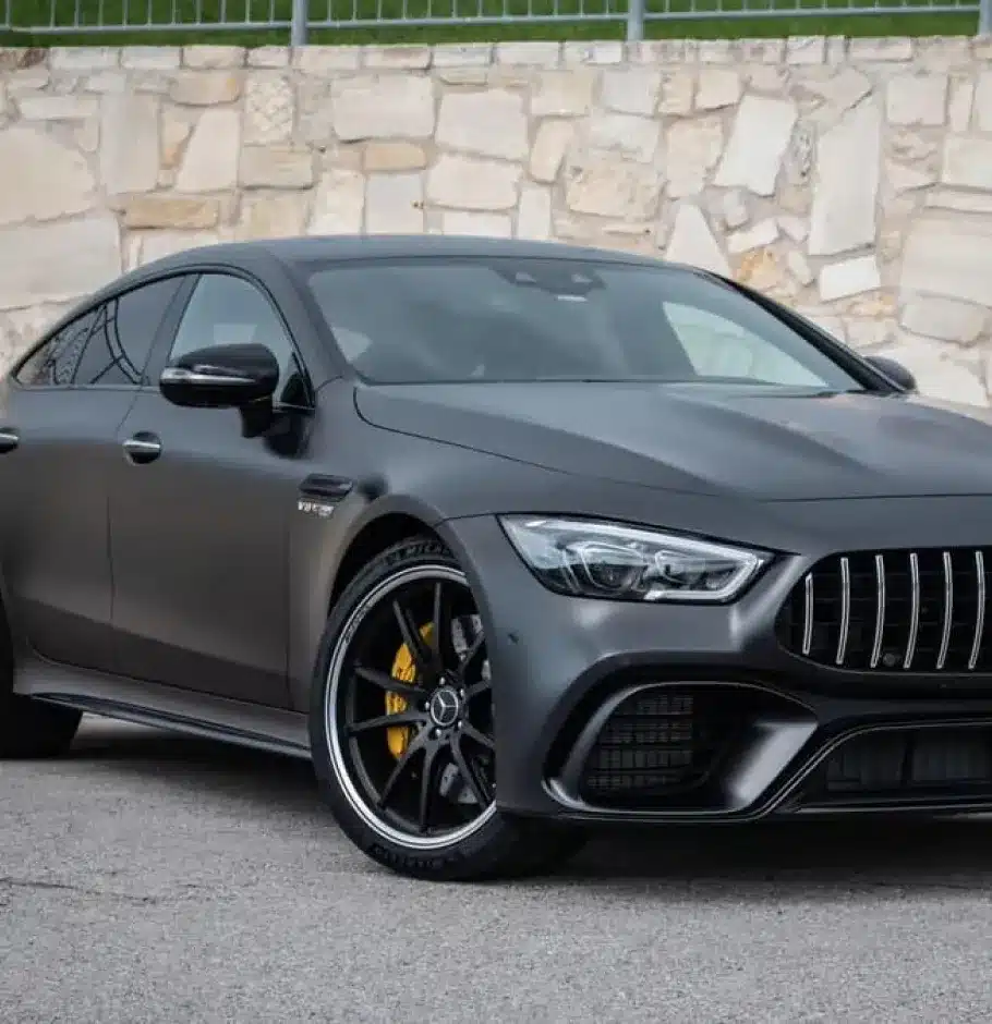 European Vehicle Repair In Monticello AR At Strokers Diesel Performance & Repair. A black Mercedes-AMG GT 4-Door Coupe is parked on a street in front of a stone wall, showcasing its sleek body, prominent grille, and yellow brake calipers behind black alloy wheels.