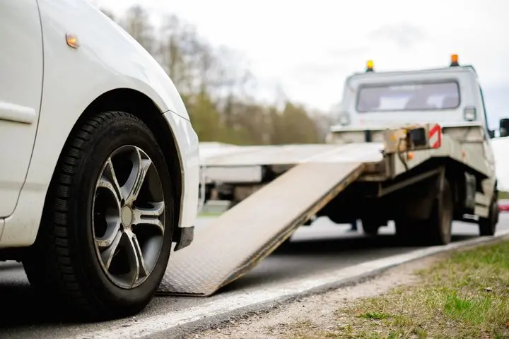 A white car is parked on the roadside, with a tow truck’s ramp extended, preparing to load the car. The background is slightly blurred, showing trees and a cloudy sky.